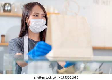 Attractive Asian Female Cafe Worker Wears Face Mask And Gloves Giving Takeaway Food Bag To Customer. 
Waitress Holding Takeout Order Standing In Coffee Shop Front Door Entrance New Normal Lifestyle