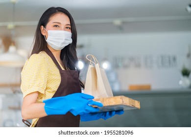 Attractive Asian Female Cafe Worker Wears Face Mask And Gloves Giving Takeaway Food Bag To Customer. 
Waitress Holding Takeout Order Standing In Coffee Shop Front Door Entrance New Normal Lifestyle