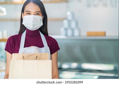 Attractive Asian Female Cafe Worker Wears Face Mask And Gloves Giving Takeaway Food Bag To Customer. 
Waitress Holding Takeout Order Standing In Coffee Shop Front Door Entrance New Normal Lifestyle