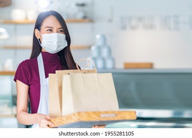 Attractive Asian Female Cafe Worker Wears Face Mask And Gloves Giving Takeaway Food Bag To Customer. 
Waitress Holding Takeout Order Standing In Coffee Shop Front Door Entrance New Normal Lifestyle