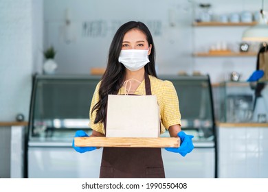 Attractive Asian Female Cafe Worker Wears Face Mask And Gloves Giving Takeaway Food Bag To Customer. 
Waitress Holding Takeout Order Standing In Coffee Shop Front Door Entrance New Normal Lifestyle