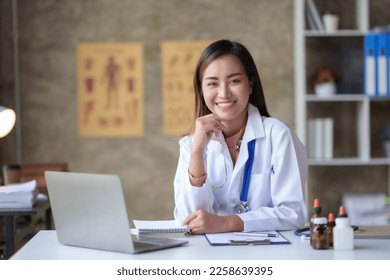 Attractive Asian female doctor checking work on desk, medicines and patient papers and using laptop computer in office room. - Powered by Shutterstock