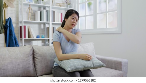 Attractive Asian Female Touching Neck And Shoulder In Pain. Young Housewife Woman Stretching Body Healthcare And Medical Painful Concept. Illness Lady Massage Back Ache Sitting On Couch Resting.