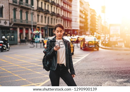 Attractive asian female tourist dressed stylish black leather jacket and black skinny jeans using connection in roaming while walking.Young charming woman enjoying leisure time at street - Imagen