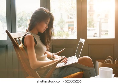 Attractive Asian Female Is Typing Messages On A Laptop While Sitting Against The Wide Windows Of A Coffee Shop. A Hipster Girl Is Working On A Portable Computer In A Cafe.