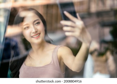 Attractive Asian Female Waving Hand To Say Hi Video Call To Her Friend By Smartphone In Cafe Restaurant Photo Shot Through Window Shop