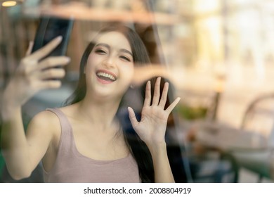 Attractive Asian Female Waving Hand To Say Hi Video Call To Her Friend By Smartphone In Cafe Restaurant Photo Shot Through Window Shop