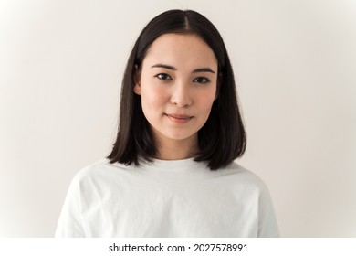 Attractive Asian Girl Wearing White T Shirt Looking Straight Into The Camera. Young Woman Posing On A White Wall Background 