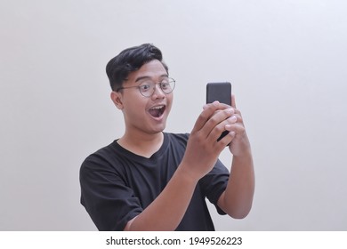 Attractive Asian man in black t-shirt wearing glasses, smiling happy while looking at his phone. Teenager holding a cellphone with arms raised. Isolated image on gray background