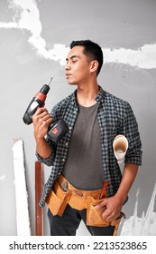 An Attractive Asian Man Blows Drill Standing With DIY Tools And Toolbelt