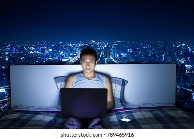 Attractive Asian Man Using Laptop Computer On His Bed At Night Doing Hard Work And Blue Light Reflection From Computer Screen