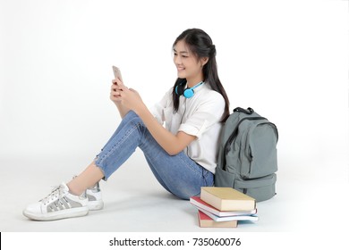 Attractive Asian Student Listening To Music From Mobile Phone With Colorful Books And Grey Bag Beside Her On White Background. Beautiful Young Girl Chat With Her Friend On Mobile Phone.