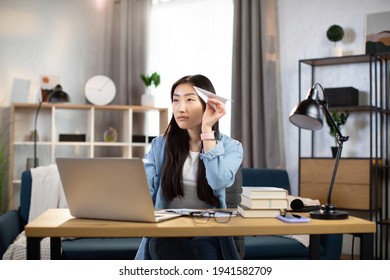 Attractive Asian Woman In Casual Outfit Sitting At Desk With Modern Laptop And Playing With Paper Plane. Female Boring At Work. Unproductive Concept.