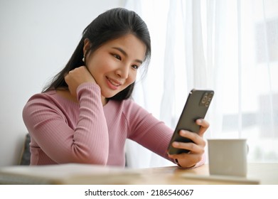 Attractive Asian Woman Chilling In The Cafe And Using Her Smartphone To Chat With Someone Or Scroll On Her Social Media. 