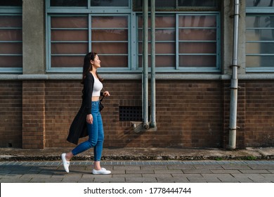 attractive asian woman with cute smile in stylish crop top in fashion blue jeans walking on street in old city near vintage red brick wall. side view full length beautiful girl relax enjoy sunshine - Shutterstock ID 1778444744