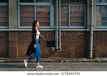 attractive asian woman with cute smile in stylish crop top in fashion blue jeans walking on street in old city near vintage red brick wall. side view full length beautiful girl relax enjoy sunshine
