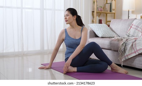 attractive asian woman gazing afar is sitting and twisting her upper body with her legs crossed after workout in a home interior at daytime.