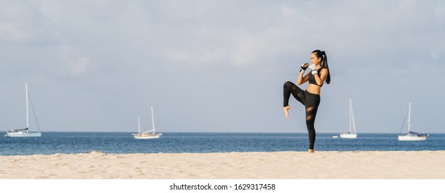 Attractive Asian woman in gloves is training on a tropical beach overlooking the sea and yachts. Thai boxing, knees, single training. Banner.