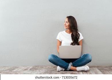 Attractive Asian Woman Holding Laptop Computer While Sitting On The Floor With Legs Crossed And Looking Away At Copy Space Isolated Over Gray Background