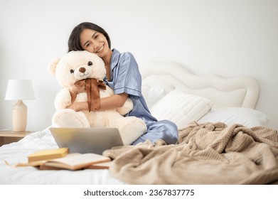 An attractive Asian woman in pajamas is hugging a teddy bear while sitting on her bed in her bedroom. Lifestyle concept