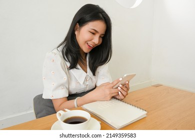 Attractive Asian Woman Relaxes In The Coffee Shop, Enjoy Reading Online Content Or Chatting With Her Friends On Smartphone.