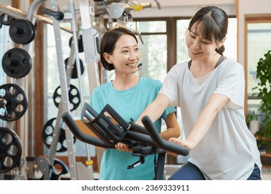 attractive asian woman using exercise bike and female trainer teaching at personal gym - Powered by Shutterstock