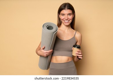 Attractive athletic woman fitness trainer or instructor in stylish sportswear standing on brown background with rolled mat and plastic cup of coffee, having rest after training lesson