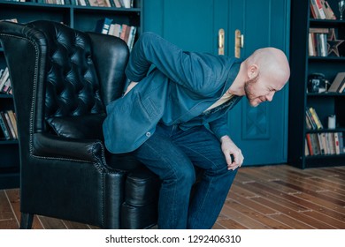 The Attractive Bald Bearded Man Is Holding Onto His Back, Trying To Get Up From His Leather Chair. Rich People Cry Too