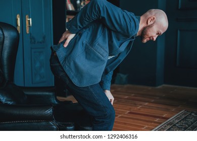 The Attractive Bald Bearded Man Is Holding Onto His Back, Trying To Get Up From His Leather Chair. Rich People Cry Too