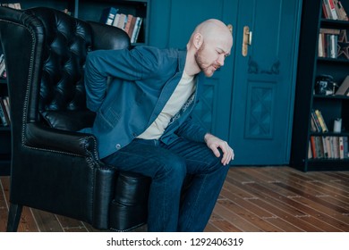 The Attractive Bald Bearded Man Is Holding Onto His Back, Trying To Get Up From His Leather Chair. Rich People Cry Too