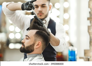 Attractive Barber With Dark Hair Wearing White Shirt, Watch And Black Gloves Doing A Haircut For Client With Scissors At Barber Shop, Lights And Mirror At Background.