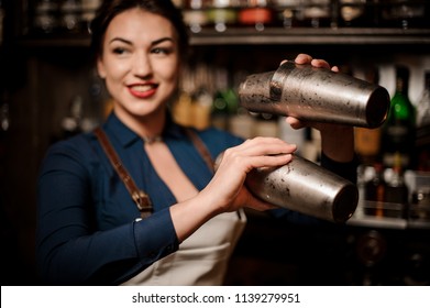 Attractive bartender girl in the white apron holding in her hands two steel cocktail shakers at the bar counter