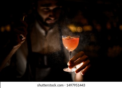 Attractive bartender serving a brown orange cocktail spraying on it in the dark on the bar counter