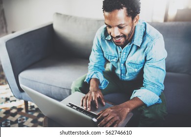Attractive Bearded African Man Spending Free Time In Sofa, Writing Email On Laptop At Modern Home.Concept Of Young People Enjoying Mobile Devices.Blurred Background.