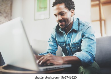 Attractive bearded African man spending free time in sofa and using laptop at modern home.Concept of young people enjoying mobile devices.Blurred background.