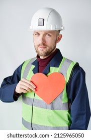 Attractive Bearded Caucasian Construction Supervisor, Foreman, Contractor Or Engineer Standing By The Wall Holding Red Heart Shape. 1st Of May, International Labour Day Concept. High Quality Image