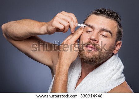 Attractive bearded guy is plucking eyebrows with concentration. He is standing and holding pincers. The hipster is looking forward seriously. Isolated