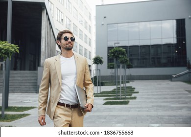 Attractive Bearded Man In Business Suit And Round Sunglasses Going To Working Meeting With Portable Laptop. Happy Businessman Standing Outdoors And Smiling.