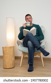 Attractive bearded man drinking a coffee while is reading a book on a modern armchair. - Shutterstock ID 1907780002