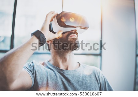 Attractive bearded man enjoyingvirtual reality glasses in modern interior design coworking studio.Home play concept.Smartphone use with VR goggles headset.Flare and sunny effect,blurred background
