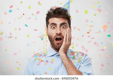 Attractive Bearded Man With Surprised Expression, Keeping His Hand On Cheek, Wearing Shirt And Party Cap, Being Shocked To Remember That He Forgot To Buy Drinks For Birthday Party. People, Celebration