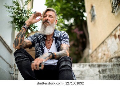 Attractive bearded man with tattoos is sitting on the stairways in the old city of athens and smoke a cigarettes