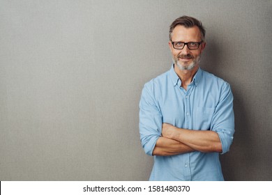 Attractive Bearded Man Wearing Glasses Standing With Folded Arms Over A Grey Studio Background With Copy Space Smiling Happily At The Camera