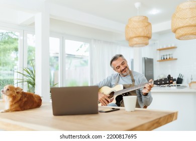 Attractive Bearded Mature Adult Male Wearing Apron Playing Acoustic Guitar, Using Laptop, Man With Dog At Home