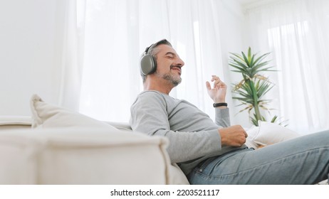 Attractive Bearded Mature Adult Man Sitting On The Couch Playing An Air Guitar And Listening To Music With Headphones, Lifestyle Concept, Relax In Living Room At Home. 