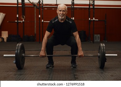 Attractive Bearded Retired Male In Black Sportswear Doing Squats And Lifting Weights In Gym. Man Smiling While Working Out, Shaping Up, Squatting With Barbell. Healthcare, Motivation And Retirement