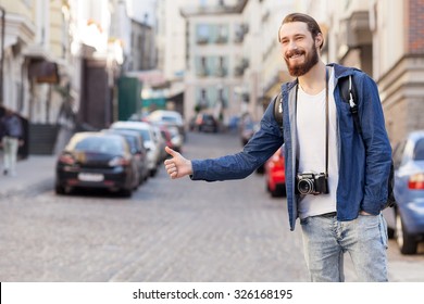 Attractive bearded tourist is hitchhiking in city. He is standing and raising hand sideways. The man is looking forward and smiling. He is carrying backpack and camera