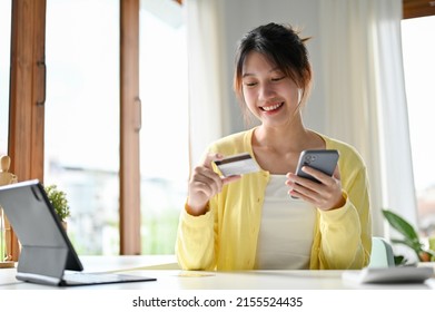 Attractive And Beautiful Asian Millennial Woman Holding A Credit Card And A Smartphone At The Table. Online Shopping, Internet Baking, Money Transfer.