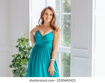 Attractive beautiful brunette girl with curls and blue blowouts in a green midi dress. Light interior room with green flowers. - Powered by Shutterstock