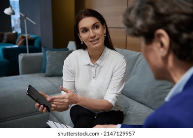 Attractive beautiful European woman showing the screen of her mobile phone to a businessman sitting in front of her in the showroom of a furniture store with a wide range of different sofas - Powered by Shutterstock